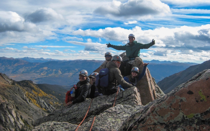 A group of students wearing safety gear rest atop a summit. One person stretches their arms out wide. There are mountains in the background.